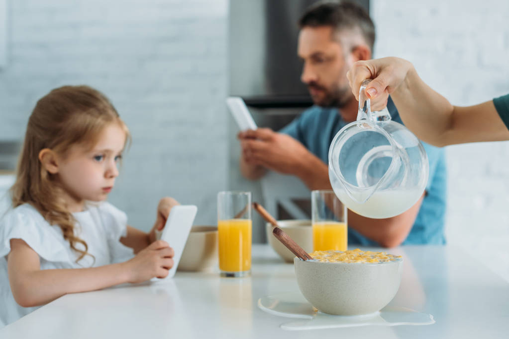 обрізаний вид на жінку, що виливає молоко в миску з пластівцями поблизу дочки і чоловіка за допомогою смартфонів
 - Фото, зображення