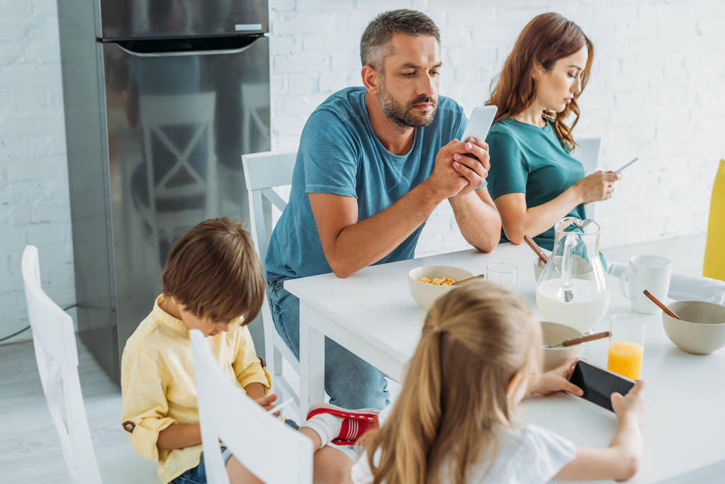 genitori con figlia e som utilizzando smartphone mentre seduti in cucina a tavola con colazione servita
 - Foto, immagini
