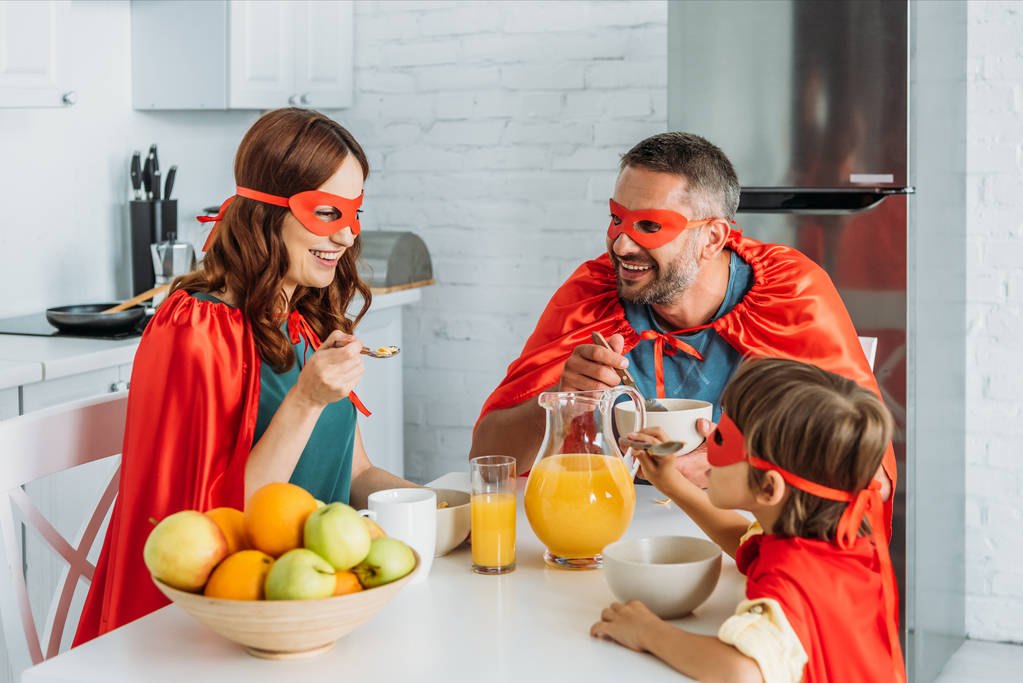 família alegre em trajes de super-heróis que tomam café da manhã na cozinha
 - Foto, Imagem