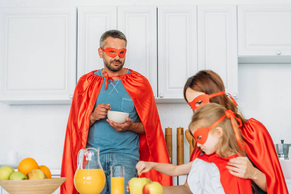 father, mother and daughter in costumes of superheroes having breakfast in kitchen  - Photo, Image