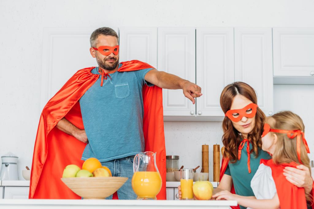 Ojciec ubrany w superhero kostium wskazując palcem na córkę i żonę siedzi na stole w kuchni w kostiumy superbohaterów - Zdjęcie, obraz
