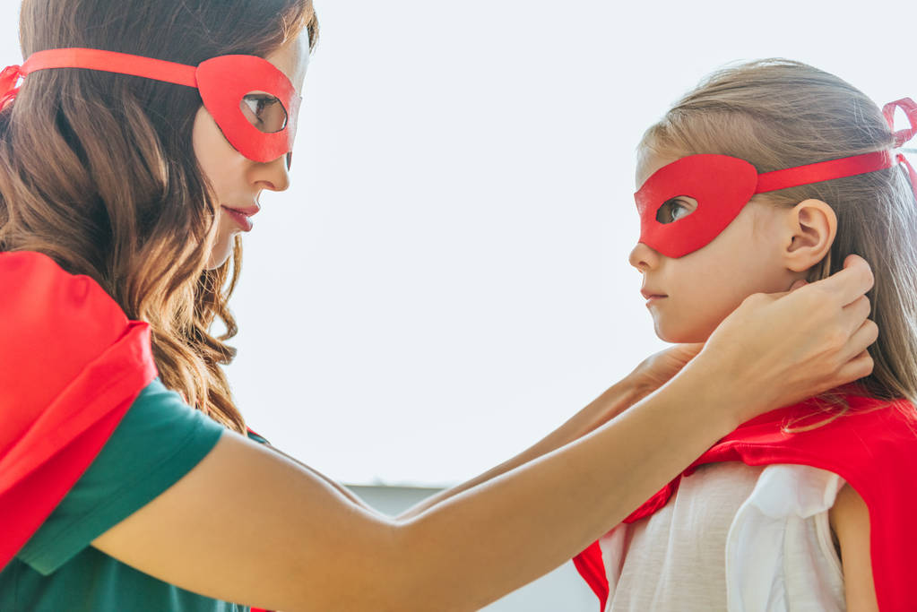 Widok z boku kobiety i urocza córka w kostiumach superbohaterów patrząc na siebie - Zdjęcie, obraz