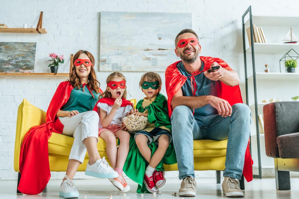familia feliz en trajes de superhéroes viendo la televisión mientras se sienta en el sofá con palomitas de maíz
 - Foto, imagen