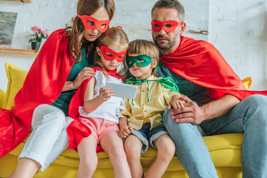 ouders met twee kinderen in kostuums van superhelden die Smartphone samen gebruiken - Foto, afbeelding