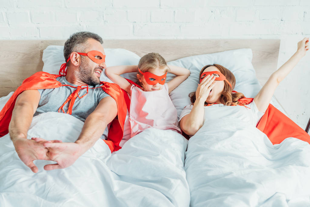 Szczęśliwa rodzina ziewanie i rozciąganie leżąc w łóżku w kostiumach superbohaterów - Zdjęcie, obraz