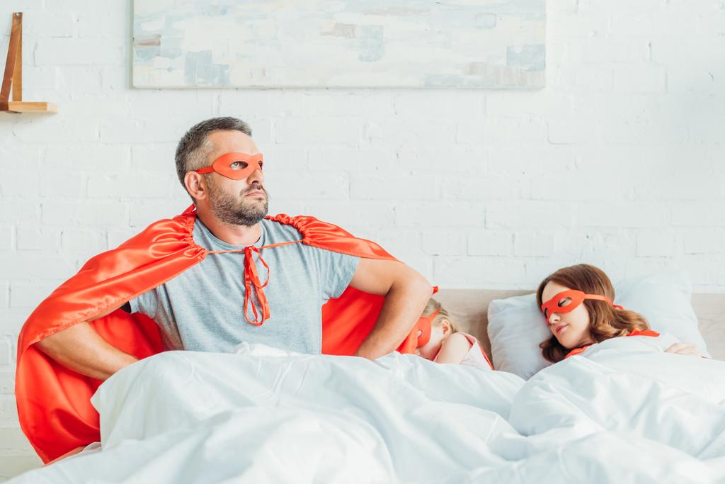 ernster Vater im Kostüm eines Superhelden sitzt im Bett neben schlafender Familie, hält die Hände an den Hüften und schaut weg - Foto, Bild