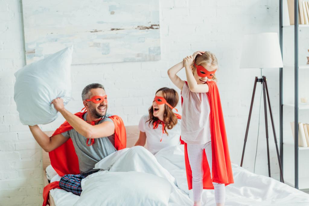 cheerful father in costume of superhero holding pillow while sitting in bed near wife and daughter - Photo, Image