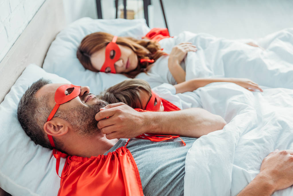 selective focus of man in superhero costume scratching cheek while lying in bed near wife and son - Photo, Image