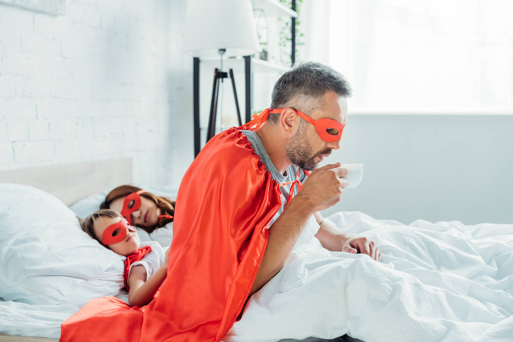 眠っている妻と息子の近くのベッドに座っている間、コーヒーを飲むスーパーヒーローの衣装を着た父 - 写真・画像