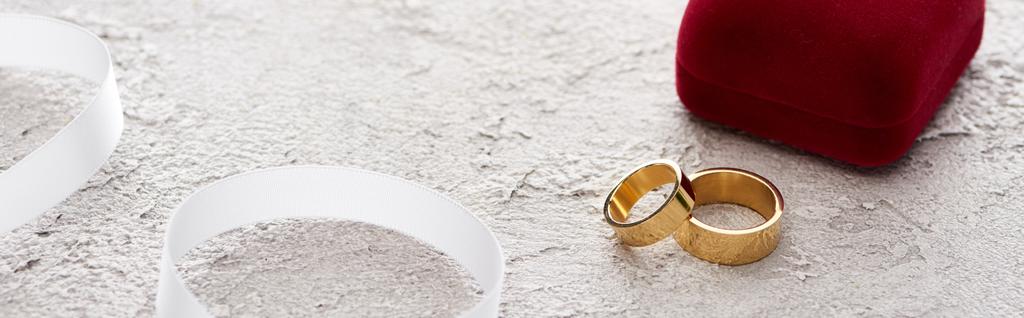 Panoramabild von goldenen Ringen in der Nähe von Geschenkschachtel und weißem Band auf strukturierter Oberfläche - Foto, Bild