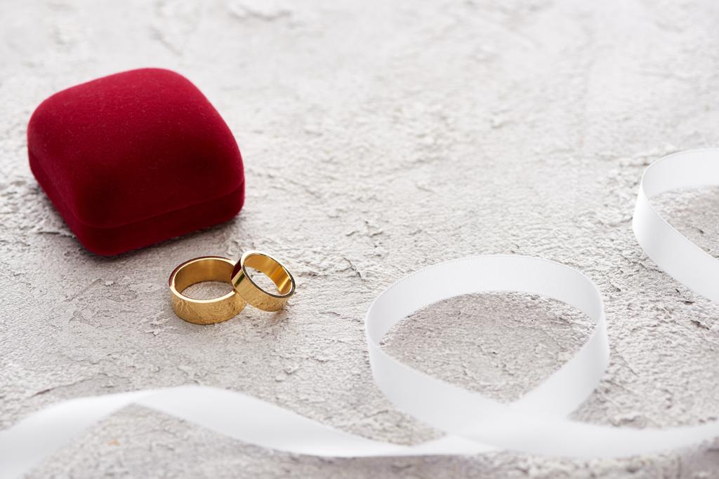 золотые кольца рядом с красной подарочной коробкой и белой лентой на текстурной поверхности
 - Фото, изображение