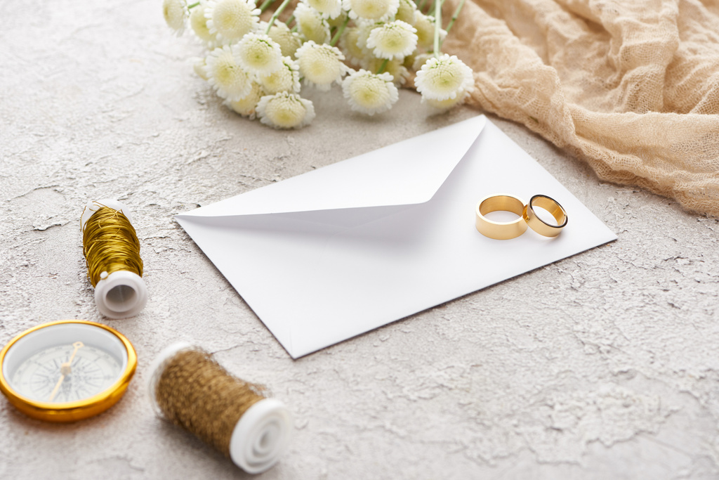 χρυσά δαχτυλίδια σε λευκό φάκελο κοντά σε μπομπίνες, χρυσάνθεμα, μπεζ σάκκα και χρυσή πυξίδα στην υφή της επιφάνειας  - Φωτογραφία, εικόνα