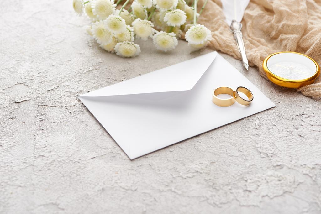 χρυσά δαχτυλίδια σε λευκό φάκελο κοντά σε χρυσάνθεμα, μπεζ σάκο, πένα και πυξίδα στην υφή της επιφάνειας  - Φωτογραφία, εικόνα