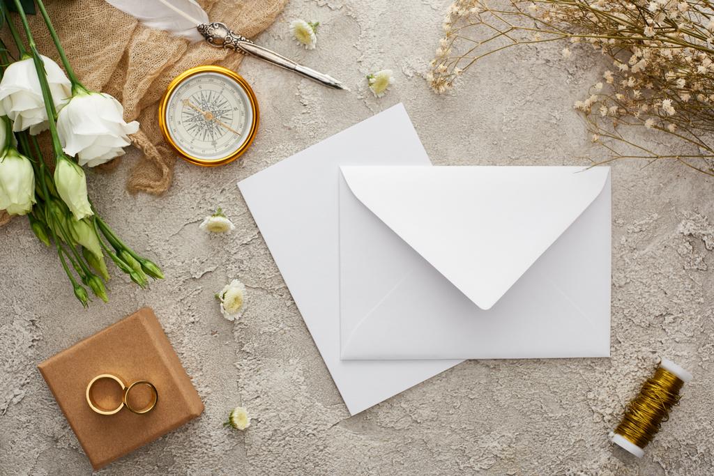 top view of white envelope and card near beige sackcloth, compass, golden rings on gift box and white flowers on grey textured surface - Photo, Image