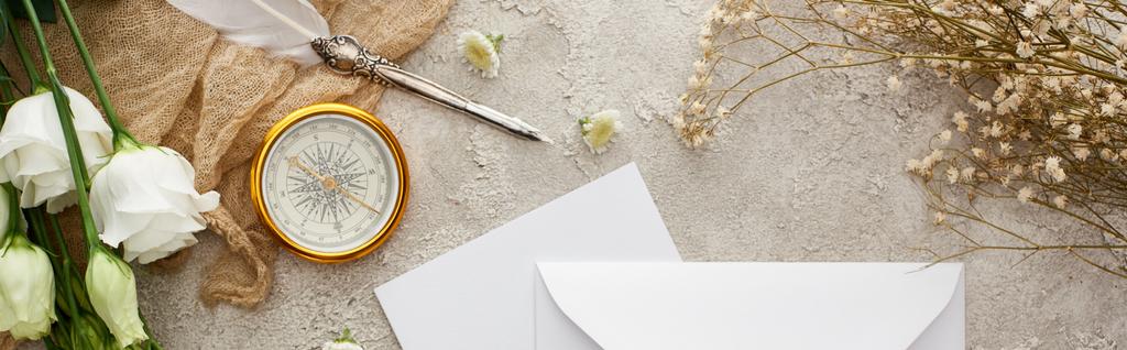 白い封筒の近くにベージュの袋の上のクイルペンのパノラマショット, 灰色のテクスチャーの表面に金色のコンパスと白い花  - 写真・画像