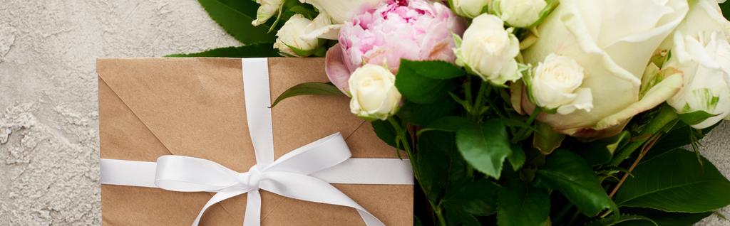 panoramische shot van envelop met wit lint in de buurt van boeket met rozen op getextureerde oppervlak - Foto, afbeelding
