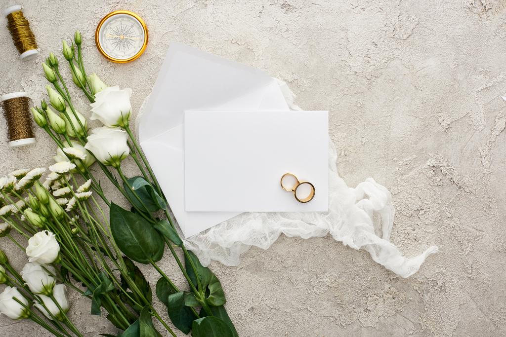 pohled na květy Eustoma u svatebních prstenců na prázdné kartě, bílé sýrové sukno, zlaté kompas a bobky na šedé, texturované ploše  - Fotografie, Obrázek