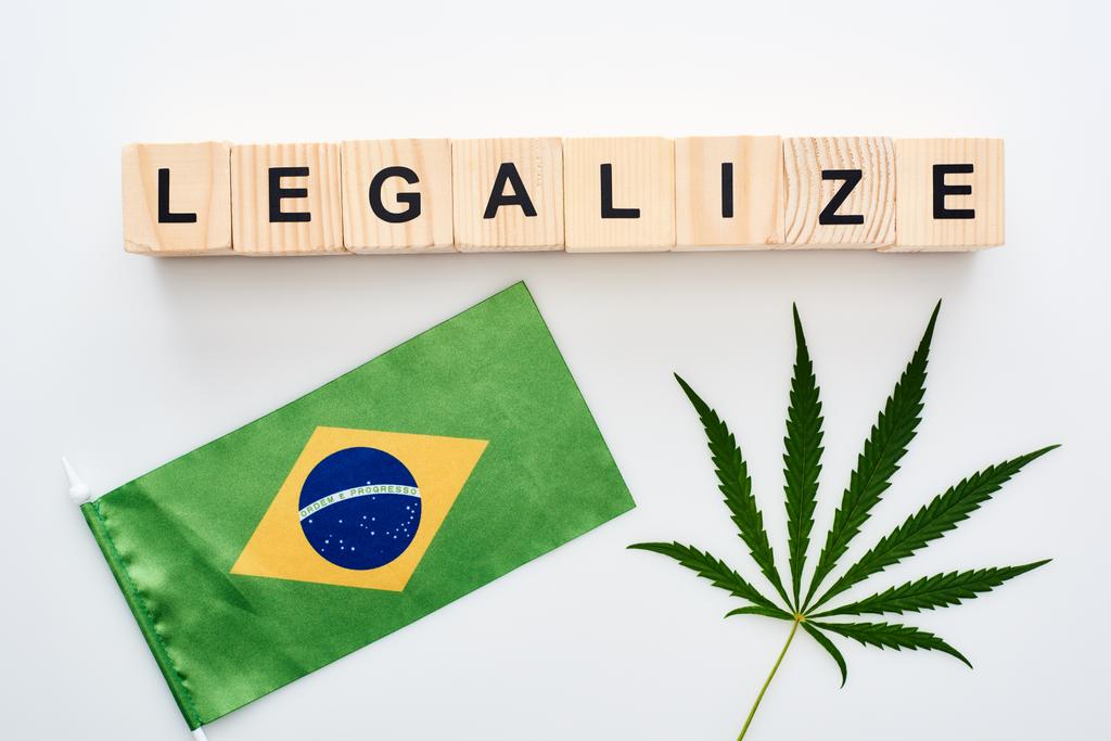 Top uitzicht van groene cannabis blad en legaliseren belettering op houten kubussen in de buurt van de vlag van Brazilië op witte achtergrond - Foto, afbeelding