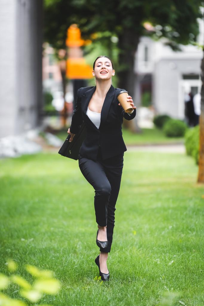 陽気で魅力的なビジネスウーマンが行くためにコーヒーを持ちながら芝生の上を走っている - 写真・画像