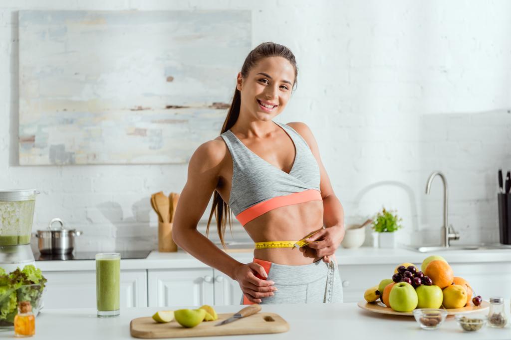 mujer feliz y deportiva sosteniendo cinta métrica mientras mide la cintura cerca de las frutas
  - Foto, imagen