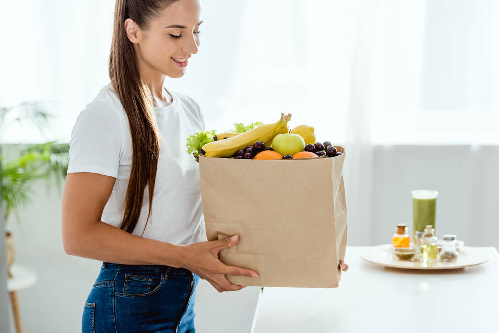 cheerful young woman looking at paper bag with fruits  - Photo, Image