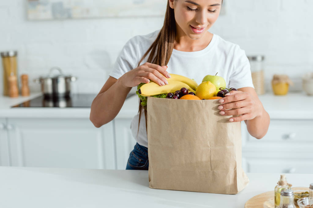 selective focus of happy girl looking at paper bag with fruits  - Photo, Image