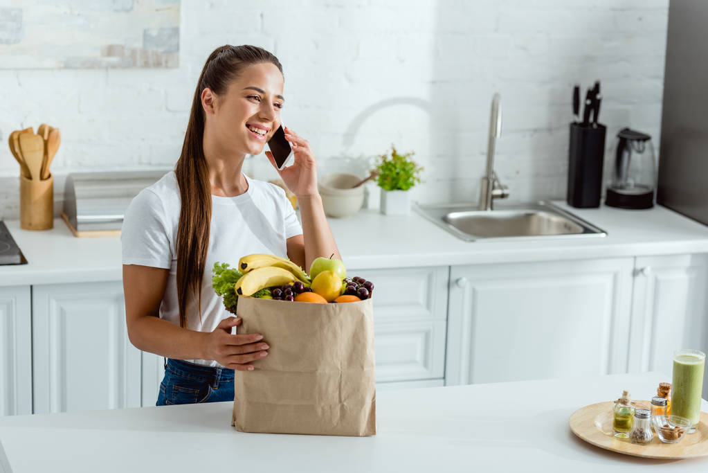 mulher feliz falando no smartphone perto de saco de papel com frutas
  - Foto, Imagem