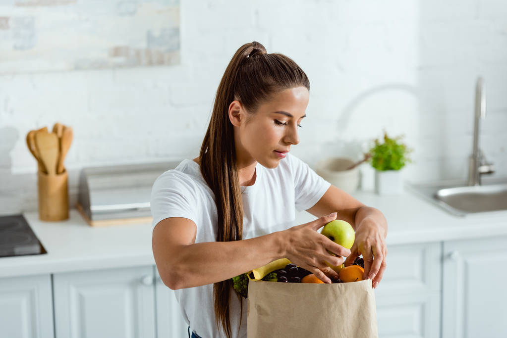 νεαρή γυναίκα κοιτάζοντας χάρτινη σακούλα και αγγίζοντας τα φρούτα  - Φωτογραφία, εικόνα