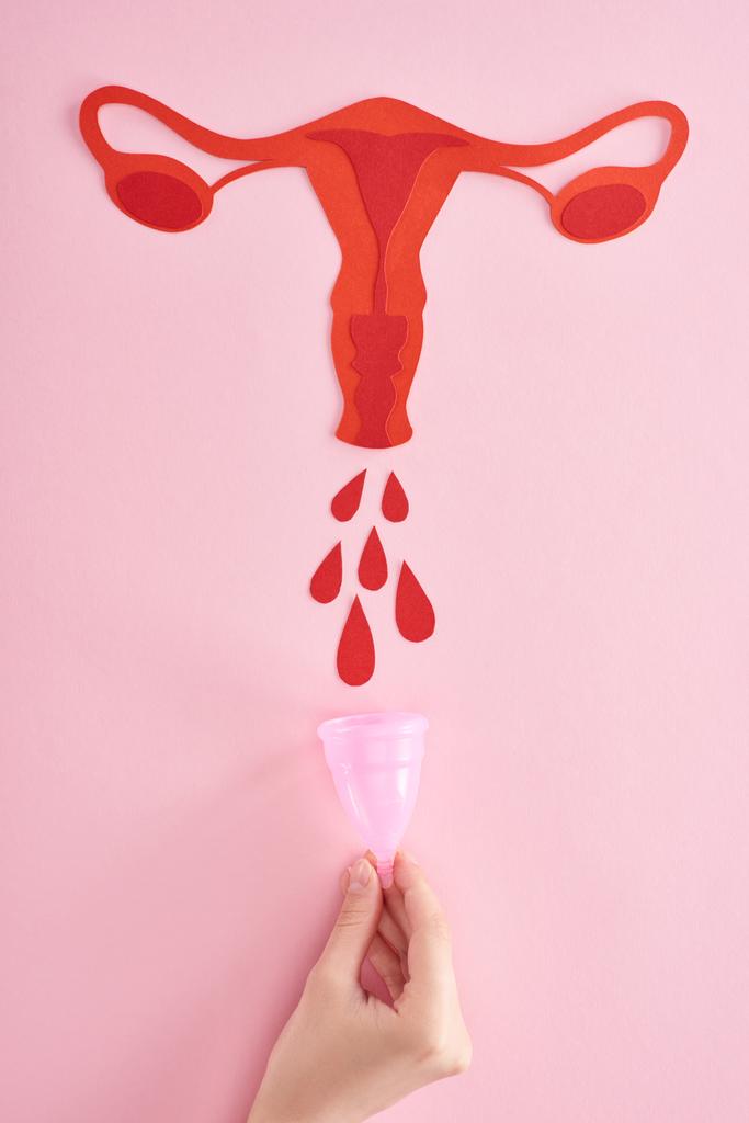 oříznutý pohled na ženu, která drží menstruální pohár v blízkosti červeného papíru, střih samičí reprodukční vnitřní orgány s krevními poklesy na růžovém pozadí - Fotografie, Obrázek