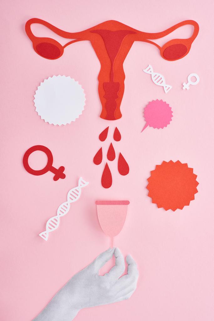 περικομμένη άποψη του χεριού κρατώντας το έμμηνο Κύπελλο κοντά σε κόκκινο χαρτί cut θηλυκό αναπαραγωγικό σύστημα με σταγόνες αίματος και άδεια φύλλα σε ροζ φόντο - Φωτογραφία, εικόνα
