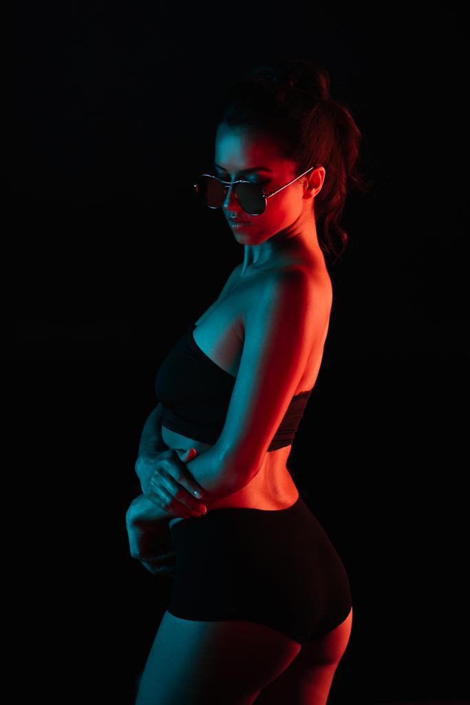mujer joven de moda en gafas de sol de pie aislado en negro
  - Foto, Imagen