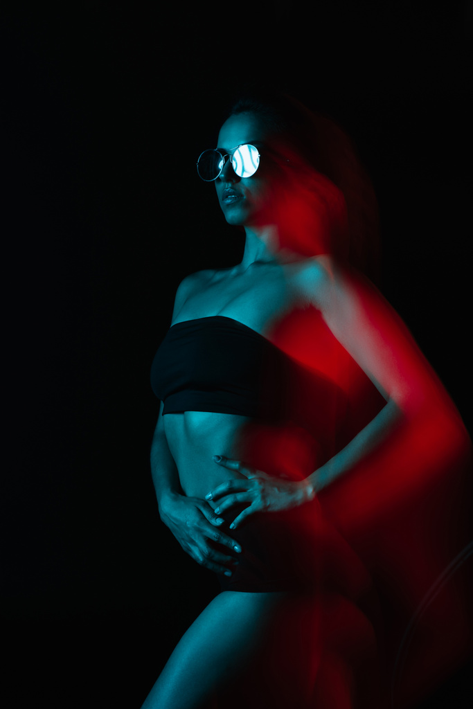 размытие движения привлекательной женщины в солнцезащитных очках, стоящей с рукой на бедре на черном
  - Фото, изображение
