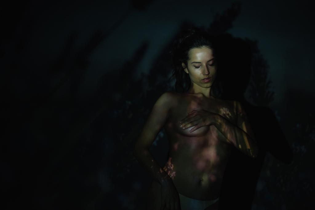 γυναίκα με σκιές στο πρόσωπο στέκεται και καλύπτοντας τα στήθη σε μαύρο  - Φωτογραφία, εικόνα