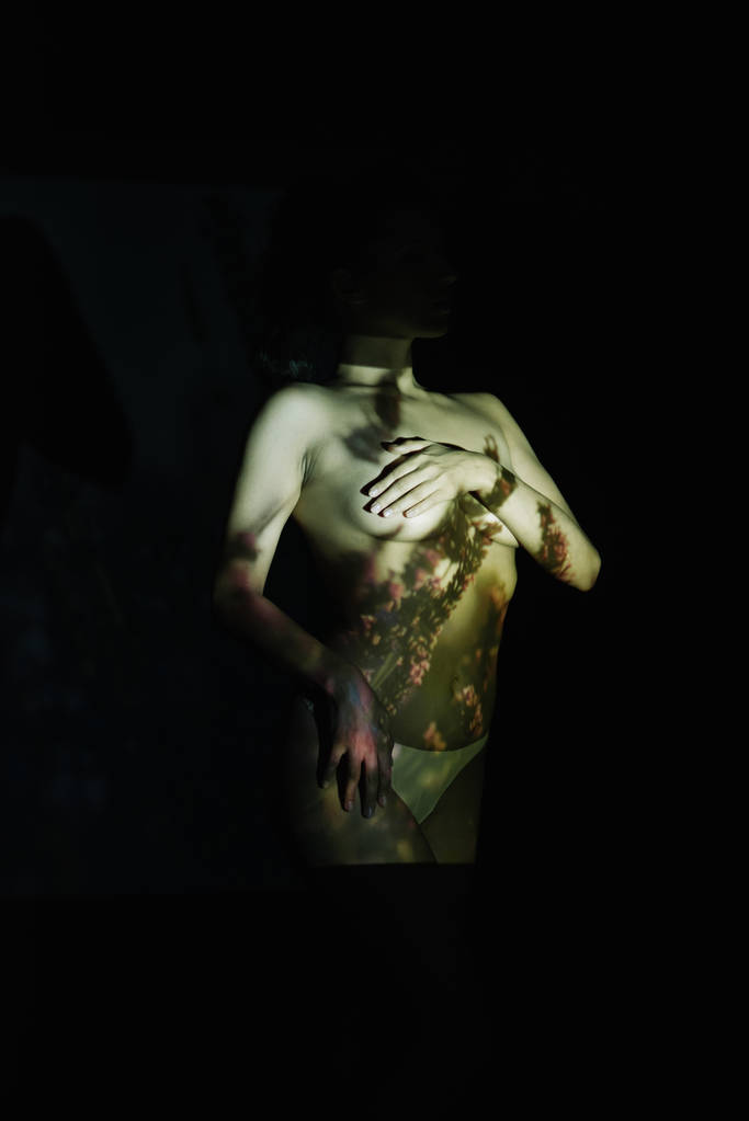 young undressed woman covering breasts while standing with hand on hip on black with shadows  - Photo, Image