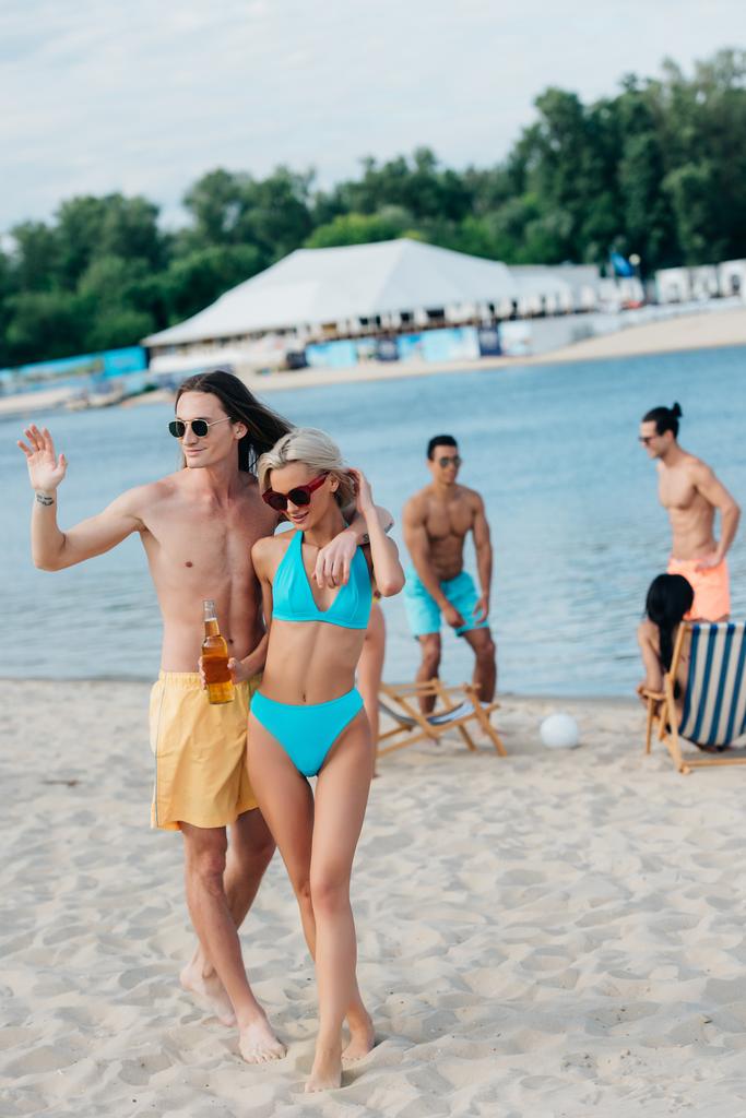Красивый молодой человек машет рукой и обнимает девушку, стоя на пляже рядом с мультикультурными друзьями
 - Фото, изображение