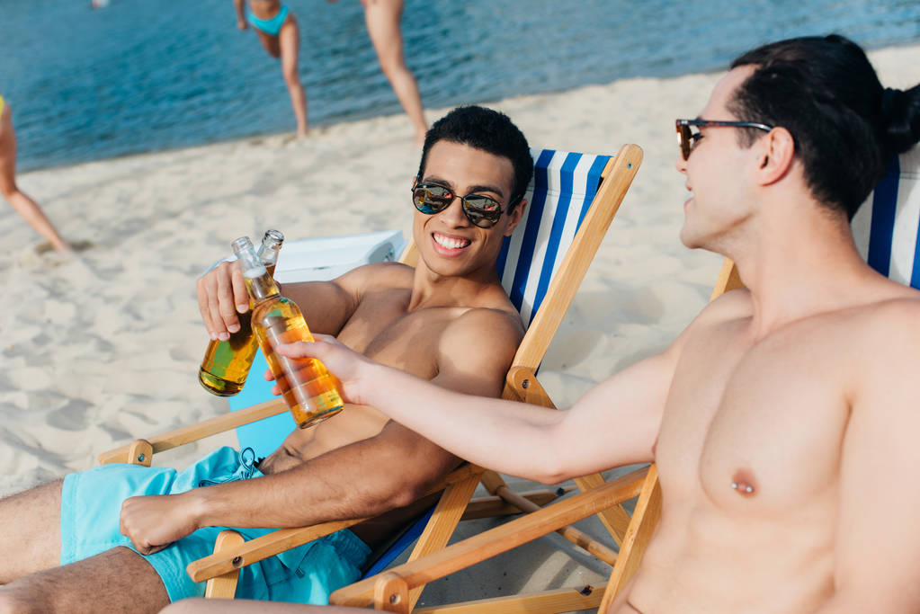 amigos multiculturales alegres tintinear botellas de cerveza mientras se sienta en chaise lounges en la playa
 - Foto, imagen