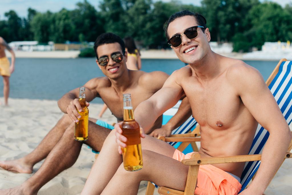 guapos amigos multiculturales sonriendo a la cámara mientras están sentados en chaise lounges y sosteniendo botellas con cerveza
 - Foto, Imagen