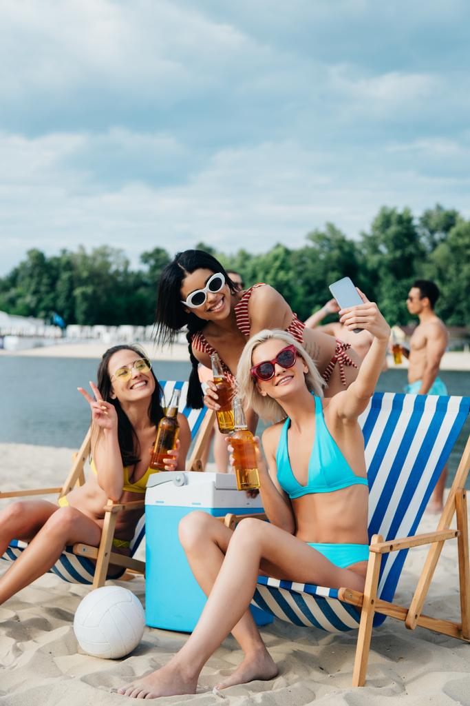 красивые мультикультурные девушки в купальниках делают селфи, выпивая пиво на пляже
 - Фото, изображение
