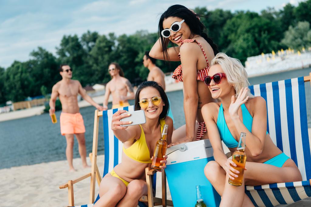 belle ragazze multiculturali prendendo selfie divertendosi sulla spiaggia vicino a bei giovani uomini
 - Foto, immagini