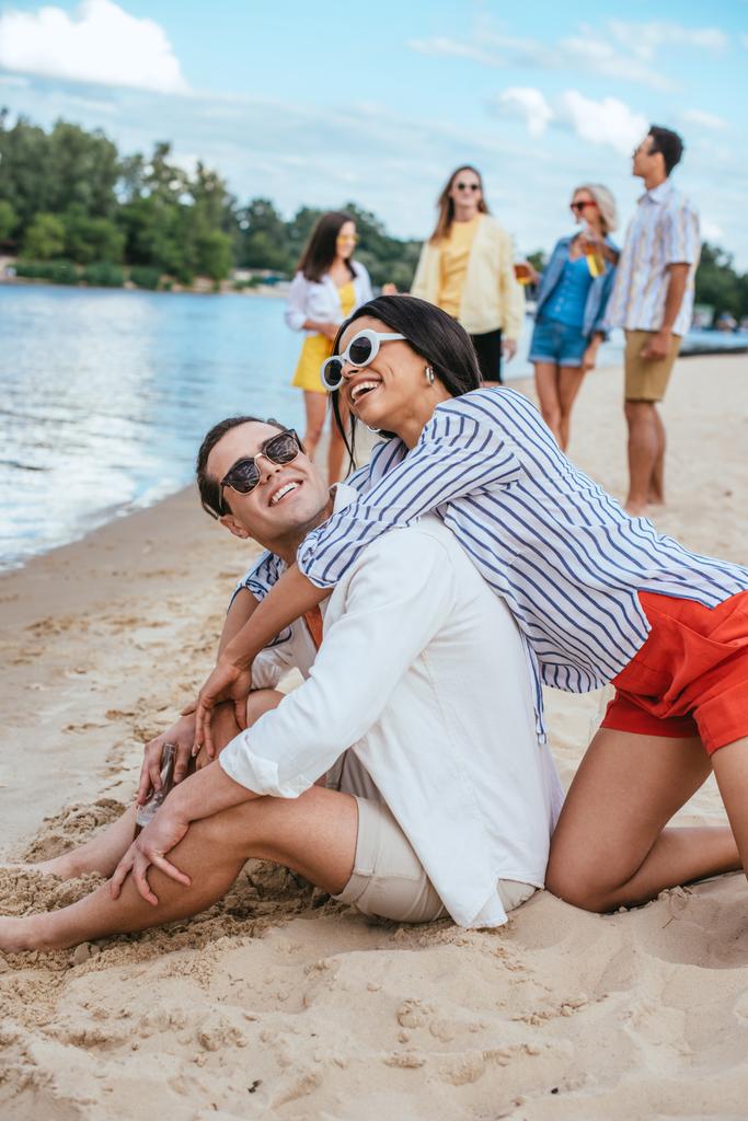 allegra coppia interrazziale divertirsi mentre seduto sulla sabbia vicino a amici multiculturali
 - Foto, immagini