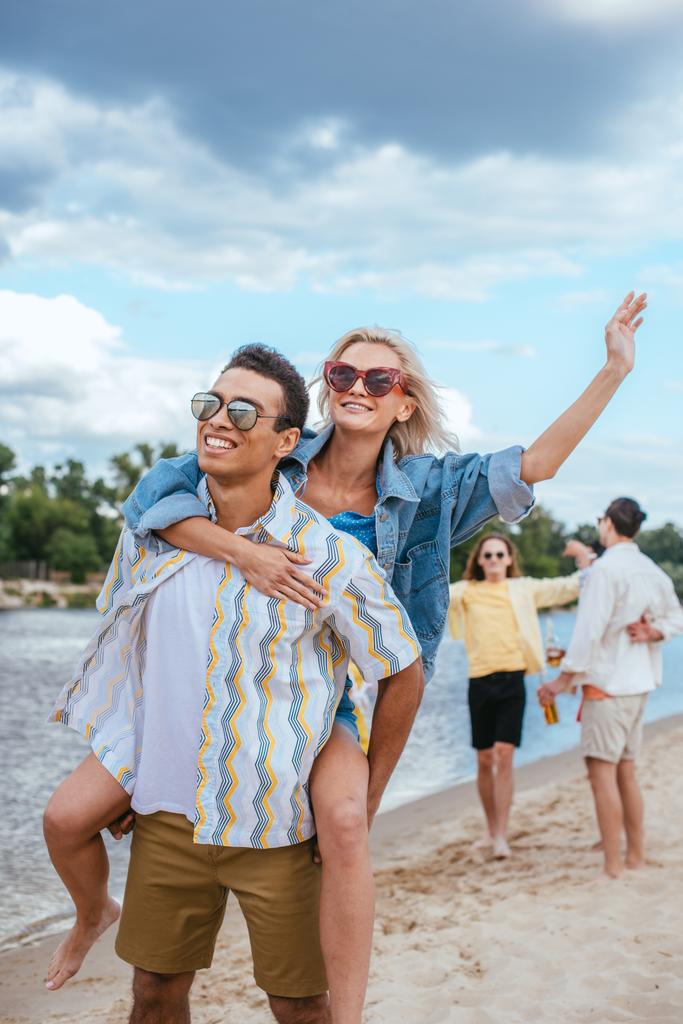 sonriente hombre de raza mixta piggybacking novia mientras camina en la playa cerca de amigos multiculturales
 - Foto, imagen