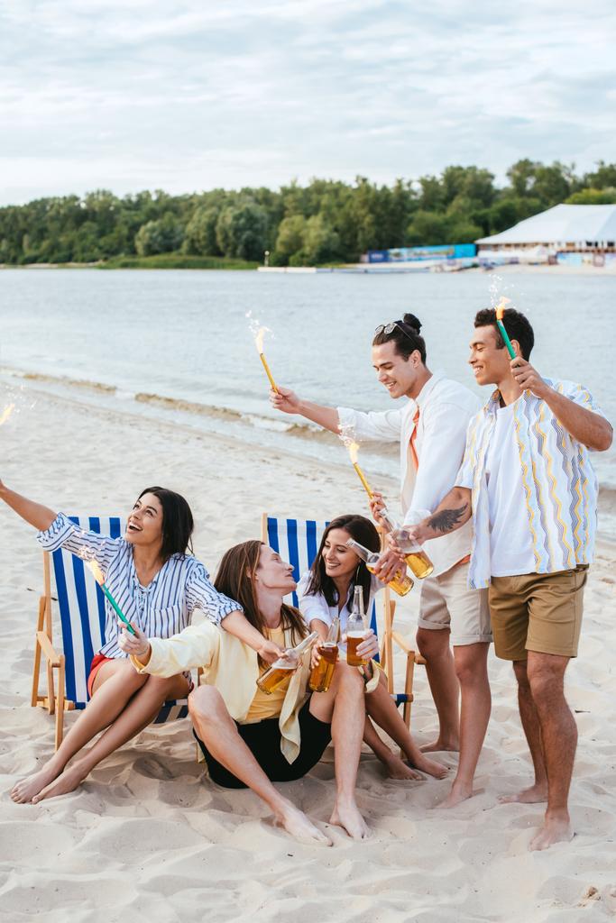 amis multiculturels joyeux tenant des bouteilles de bière et des étincelles tout en s'amusant sur la plage
 - Photo, image