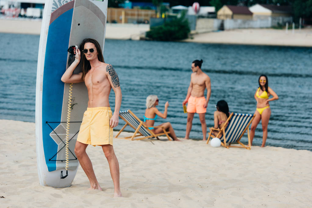 csinos fiatalember nézett kamerával, miközben állandó közelében szörfdeszka a strandon - Fotó, kép