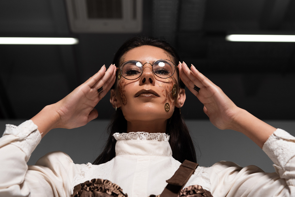 low angle view of steampunk woman touching glasses and looking at camera - Photo, Image
