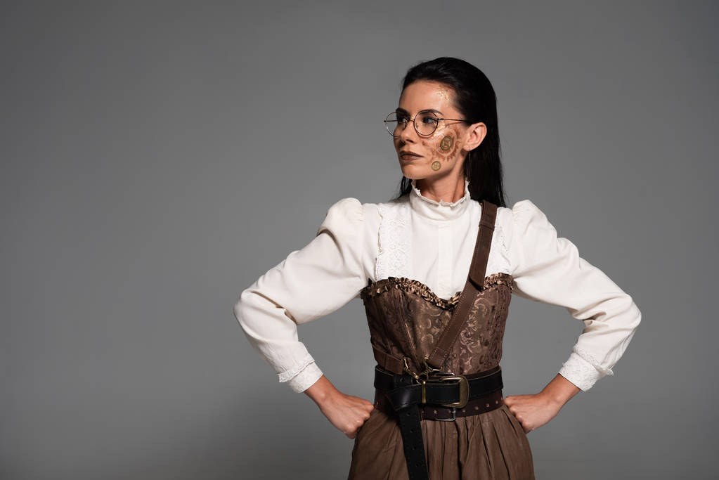 nachdenkliche Steampunk-Frau mit Brille, die Hände auf den Hüften isoliert auf grau - Foto, Bild