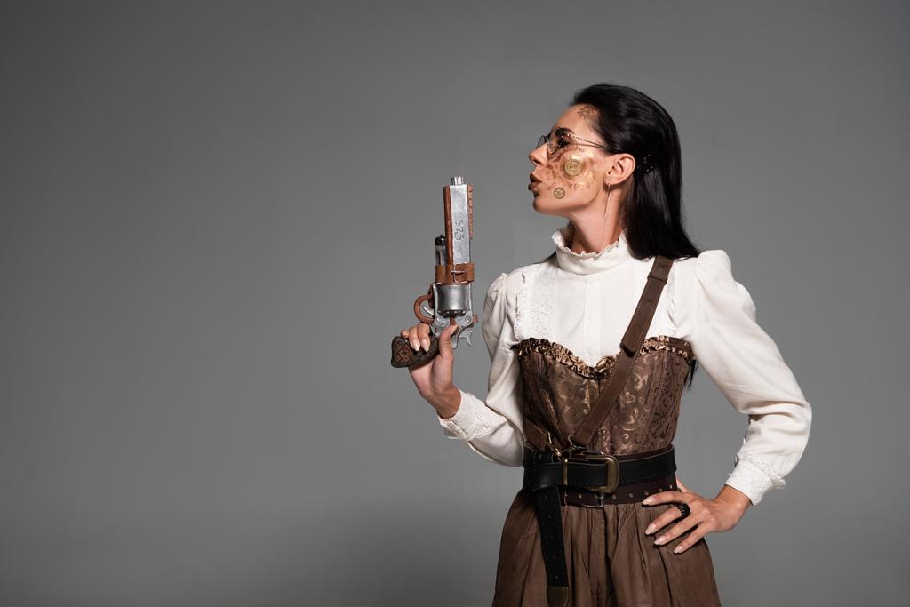 kendine güvenen steampunk kadın kalça el ile ayakta ve gri izole tabanca tutarak - Fotoğraf, Görsel