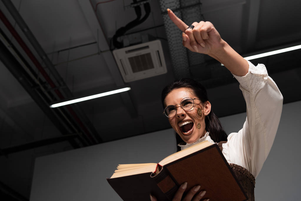 vista inferior de la mujer steampunk enojado sosteniendo libro abierto, gritando y señalando con el dedo
 - Foto, Imagen
