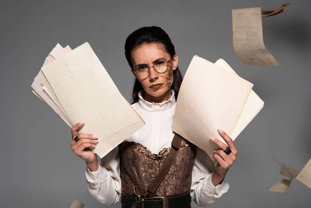 displeased steampunk woman in glasses holding documents and looking at camera on grey - Photo, Image