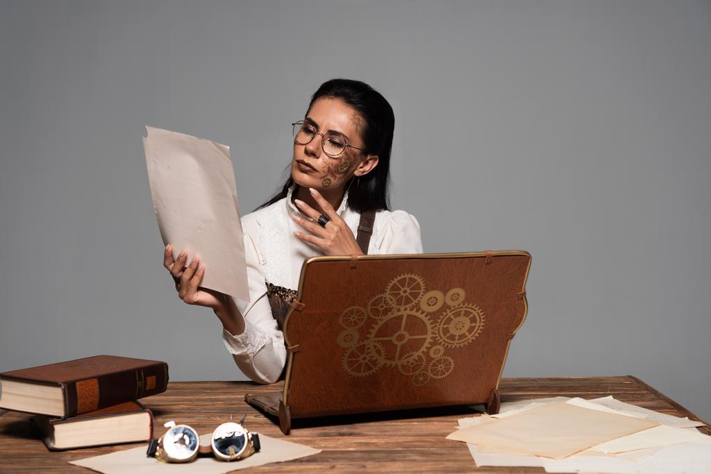 mulher steampunk pensativo em óculos com documentos e laptop vintage no local de trabalho isolado em cinza
 - Foto, Imagem