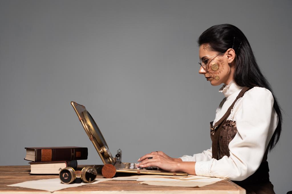 Seitenansicht der fokussierten attraktiven Steampunk-Frau mit Laptop am Arbeitsplatz isoliert auf grau - Foto, Bild